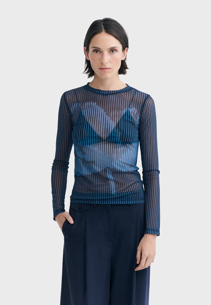 Femme aux cheveux courts et foncés portant un haut transparent bleu marine à manches longues avec des rayures verticales et un pantalon noir, debout devant un fond uni.