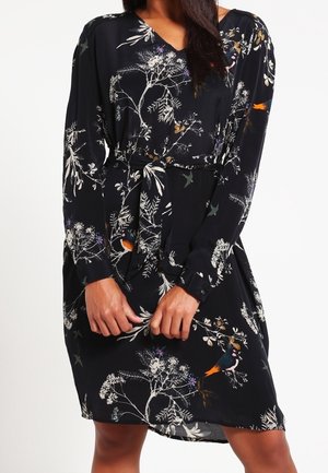 Žena v černých šatech s dlouhým rukávem s bílými květinovými a barevnými ptáčky, stažených v pase, ruce sepnuté před sebou.