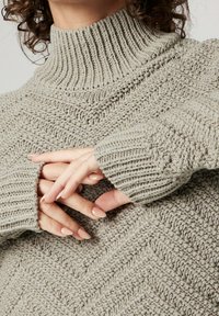 Hellgrauer Strickpullover mit hohem, geripptem Kragen und strukturiertem Chevron-Muster. Lange Ärmel mit gerippten Bündchen.