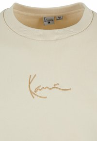 Kremfarget hettegenser med ribbet krage, med en gylden brodert logo "Kanu" på forsiden. Myk, glatt tekstur.
