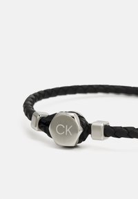 Svart flätad läderarmband med en silverfärgad sexhörningstapp som har en ingraverad "CK"-logotyp, accentuerad av släta metalliska inslag.