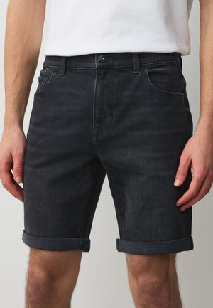 Man draagt donkere denim opgerolde shorts en een wit t-shirt, te zien van taille tot halverwege het bovenbeen tegen een effen achtergrond.