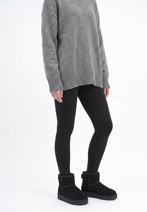 Personne portant un pull gris oversize, des leggings noirs et des bottines noires doublées de fourrure, debout devant un fond blanc.