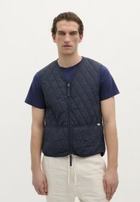 Vest quiltat de culoare bleumarin cu fermoar frontal, decolteu rotund și două buzunare frontale. Purat peste un tricou bleumarin ajustat și pantaloni de culoare deschisă.