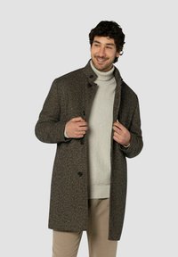 Brauner Fischgrätmantel mit hohem Kragen, schwarzen Knöpfen und lockerer Passform, getragen über einem hellgrauen Rollkragenpullover.