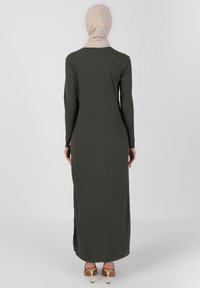 Longue robe moulante côtelée de couleur vert olive avec manches longues, fentes latérales et coupe ajustée. Portée avec des chaussures de couleur claire.