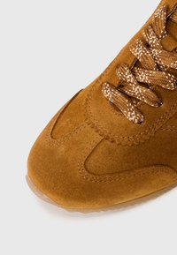 Bruine suede sneaker met patronen in de veters en gedetailleerde stiksel op een lichte achtergrond, waarbij de voorkant en zijkanten worden getoond.