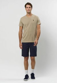 Tricou bej cu decolteu în V, asortat cu pantaloni scurți bleumarin, având un mic logo pe piept. Șosete albe și teniși bleumarin completează ținuta.