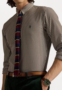 Prugasta smeđa i bijela košulja sa gumbima, s malim zelenim logom, uparena s dušicom u više boja, smeđim pojasom i zelenim satom.