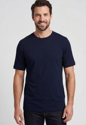 Lächelnder Mann mit kurzen dunklen Haaren und Bart, der ein schlichtes marineblaues Kurzarm-T-Shirt und dunkle Jeans vor hellem Hintergrund trägt.