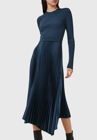 Pitkähihainen, ribattu neulemekko syvässä sinisessä, jossa on pleated-hame, epäsymmetrinen helma ja istuva yläosa. Sujuva tekstuuri.