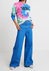 Farverig tie-dye langærmet skjorte i pink, blå og grøn med "VANS" logo. Kombineret med vide blå denim jeans og hvide sneakers.