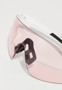 Lunettes de sport avec verres teintés roses. Monture blanche avec accents noirs et plaquettes de nez noires ajustables. Équipées de protections latérales intégrées.