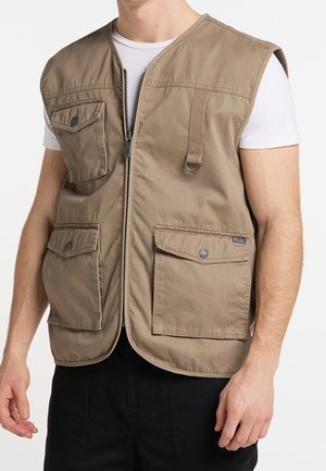 Mężczyzna noszący beżową kamizelkę roboczą z czterema przednimi kieszeniami na klapy i zamkiem błyskawicznym, na białej koszuli z krótkim rękawem i czarnych spodniach.