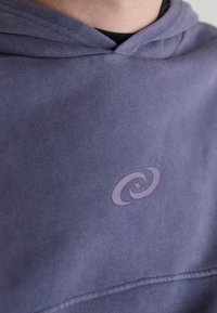 Lilla hettegenser laget av bomullsblanding, med et tonalt logo-design i en litt lysere nyanse. Inkluderer snor og teksturert stoff.