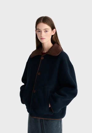 Junge Frau mit langen braunen Haaren trägt eine dunkelblaue Fleecejacke mit großem braunem Kragen und die Hände in den Taschen vor einem schlichten Hintergrund.