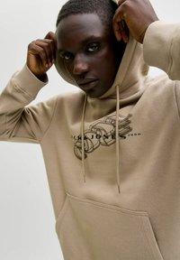 Młody mężczyzna poprawiający kaptur beżowego hoodie z czarnym abstrakcyjnym napisem "CORE" oraz "JACK & JONES 1990" na jednolitym tle.