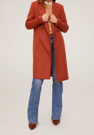 Femme portant un long manteau orange brûlé, un pull couleur chameau, un jean bleu clair et des chaussures en cuir marron pointues, debout devant un fond uni.