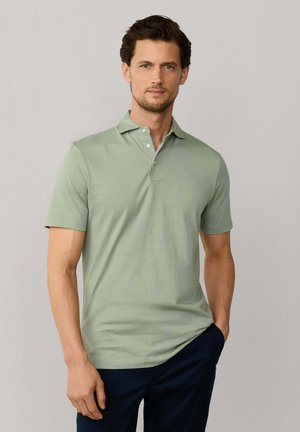 Man met een lichtgroen poloshirt met korte mouwen en een donkere broek, staand met één hand in de zak tegen een effen achtergrond.