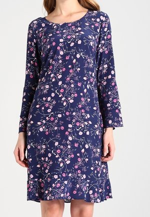 Femme portant une robe bleu marine à manches longues avec des motifs floraux roses et blancs et un ourlet à volants.