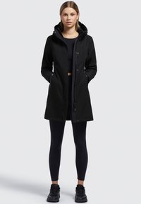 Zwarte waterdichte jas met capuchon, knopen aan de voorkant en zijzakken. Glad oppervlak, middellange stijl, gedragen met zwarte leggings en schoenen.