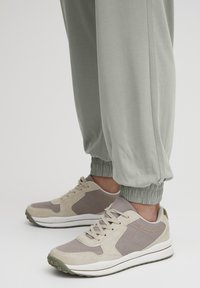 Idrottsskor i beige och grått med strukturerad mocka och mesh-övre, vit mellansula och grön gummisula. Elastiska muddar på grå joggers.