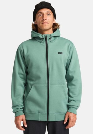 Grüne Kapuzenjacke aus weichem Stoff, mit durchgehendem Reißverschluss, zwei Fronttaschen und einem kleinen Logopatch auf der Brust.