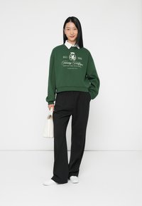 Felpa verde con logo e testo bianchi, abbinata a pantaloni neri a gamba larga e scarpe bianche. Il modello tiene una piccola borsa bianca.