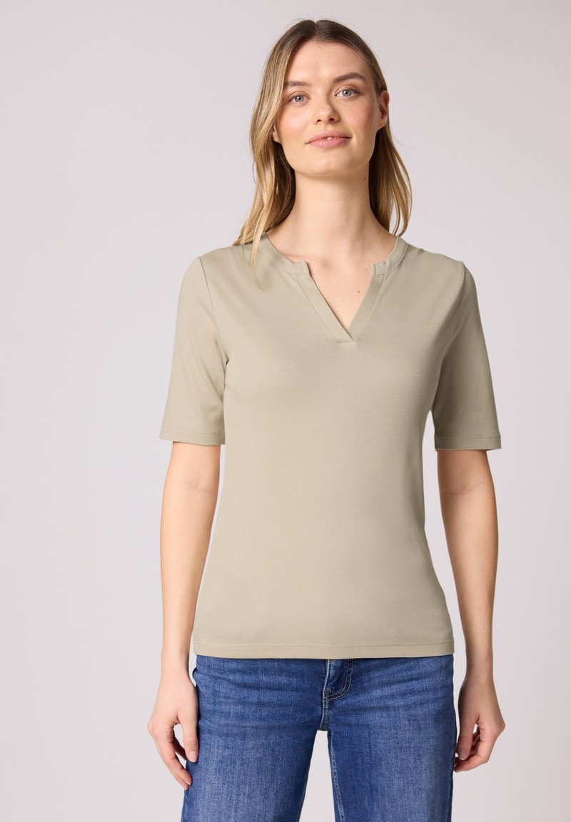 Femme portant un haut beige à manches courtes et col en V, ainsi qu'un jean bleu, debout devant un fond uni gris clair.