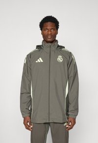 Grøn jakke med hætte, prydet med lysegrønne detaljer og Adidas-branding. Lavet af letvægtsmateriale, har den lynlås-lukning og elastiske manchetter.