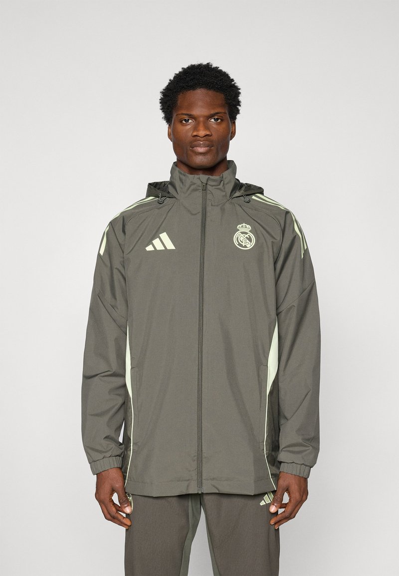 Grøn jakke med hætte, prydet med lysegrønne detaljer og Adidas-branding. Lavet af letvægtsmateriale, har den lynlås-lukning og elastiske manchetter.