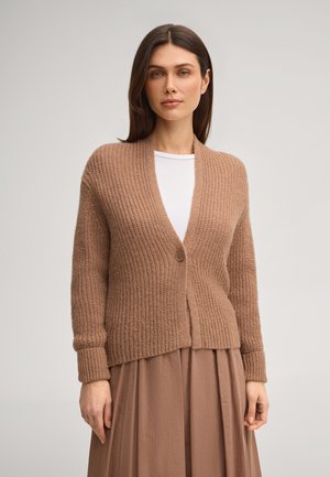 Frau mit glattem braunem Haar, die eine beige gestrickte, geknöpfte Strickjacke über einem weißen Hemd und einen braunen plissierten Rock trägt, vor einem schlichten Hintergrund stehend.