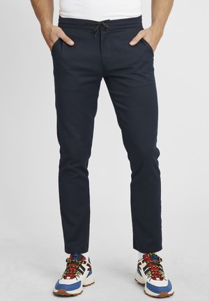 Navy blue jogger-style pants with an elastic waistband and drawstring. Smooth fabric, slim fit, pockets on both sides. Worn with colorful sneakers.