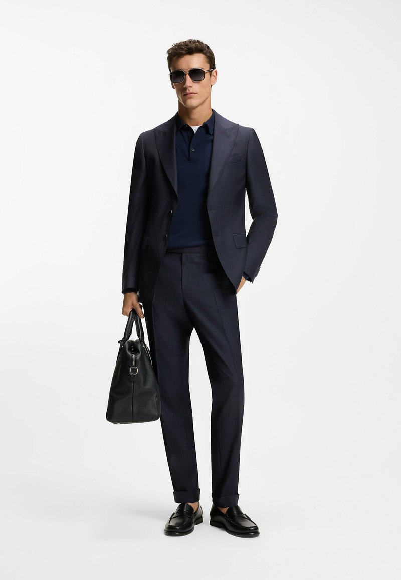 Marineblau gestreifter Anzug mit kurzärmligem Hemd, schwarze Sonnenbrille und schwarze Ledertasche. Schuhe sind schwarze Loafers.