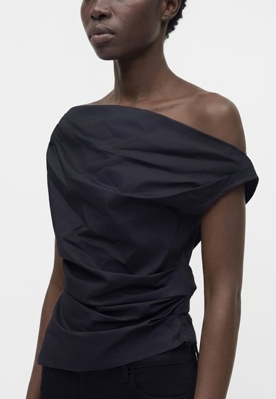 Femme portant un haut noir à épaules dénudées froncé avec un pantalon noir ajusté, debout devant un fond clair uni.