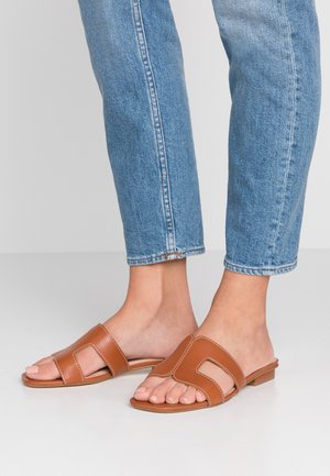 Sandales en cuir marron à enfiler avec un design à découpe, semelle plate et couture blanche, associées à un jean bleu clair retroussé au bas.
