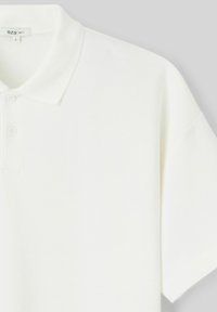 Chemise polo blanche avec un col classique, trois boutons, des manches courtes et une texture lisse et légère. Étiquette visible, taille L.