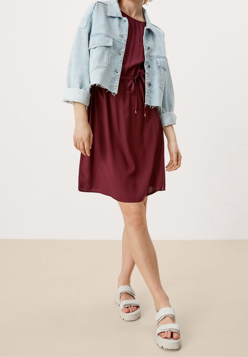 Femme portant une robe bordeaux longueur genou avec une taille nouée, une veste courte en jean clair et des sandales blanches à semelles épaisses.