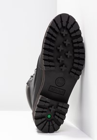 Botte en cuir noir avec semelle en caoutchouc texturée, présentant des crampons profonds pour l'adhérence, un logo et un accent vert au bas.