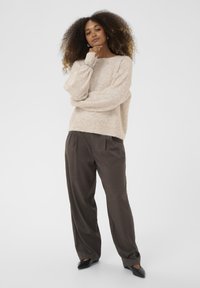 Bežani, teksturirani pulover s širokim rukavima, uparen s smešnim, visokim, plisiranim hlačama i crnim cipelama na špic.