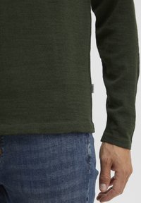 Pull en tricot vert à manches longues, avec un ourlet droit et une petite étiquette sur le côté, associé à un jean en denim bleu.