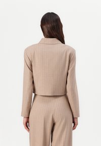 Beskyttende beige blazer med vertikale hvite striper. Strømlinjeformet design med utskårede revers og lange ermer, laget av glatt stoff.