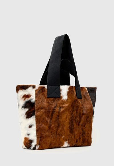 Sac fourre-tout avec motif en cuir de vache marron, blanc et noir et larges anses en tissu noir sur fond gris clair.