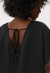 Zwarte blouse met een V-vormige achterkant en een striksluiting, wijde mouwen en een soepele textuur. Opvallend detail is een kleine knoopaccent.