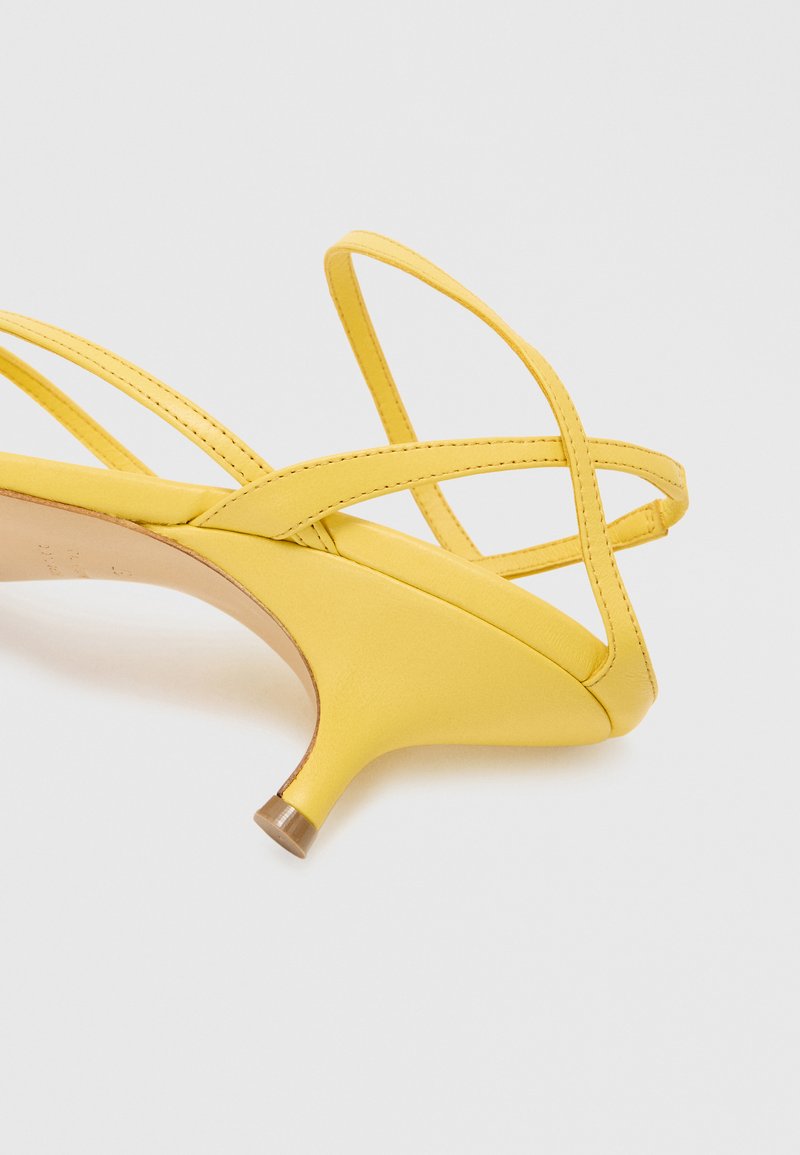 Sandal de tacón amarillo con correas entrelazadas, punta afilada y un tacón delgado con un acento metálico en la base. Textura suave.