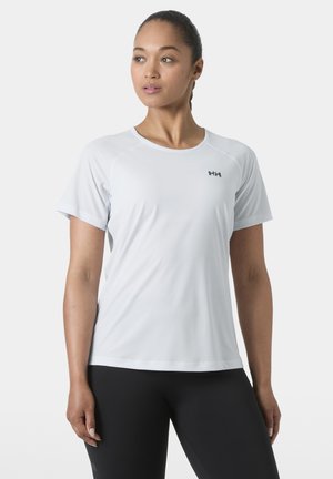 Kobieta ubrana w biały sportowy T-shirt z krótkim rękawem z logo Helly Hansen, w połączeniu z czarnymi legginsami, stojąca na plain jasnym tle.