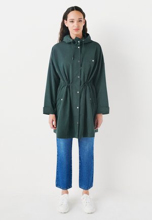 Veste verte légère avec une taille à cordon de serrage, fermeture à boutons sur toute la longueur et capuche. Comprend deux poches avant et une coupe décontractée.