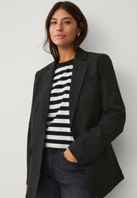 Blazer noir avec des épaules structurées, fermeture à bouton unique et revers crantés, porté sur une chemise à rayures noires et blanches.
