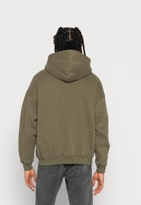 Sudadera verde oliva con una capucha grande, hombros caídos y puños acanalados. La textura de la tela parece suave; presenta un diseño sencillo y relajado.