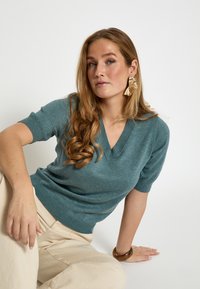 Maglione lavorato a maglia con scollo a V color verde acqua, a maniche corte, realizzato in materiale morbido. Il modello indossa pantaloni a gamba larga color crema e un bracciale spesso.